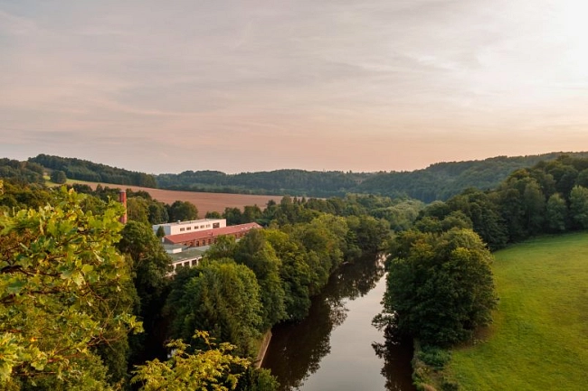 Das Bild zeigt die Zschopau in Waldheim. Der Fluss verläuft geschwungen durch ein grünes Tal und spiegelt den leicht bewölkten Himmel wider. Beide Ufer sind dicht mit Bäumen bewachsen. Links im Bild steht ein größeres Gebäude mit hellem Dach, das teilweise von Bäumen umgeben ist. Im Hintergrund sind sanfte Hügel und Felder zu sehen. Das Licht wirkt warm, wie am späten Nachmittag oder frühen Abend. Die Szene vermittelt eine ruhige, naturnahe Atmosphäre.
