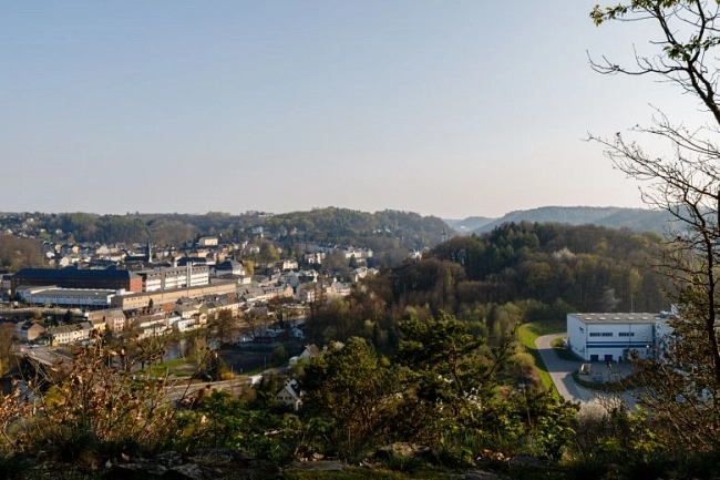 Das Bild zeigt einen weiten Blick über die Stadt Waldheim aus erhöhter Perspektive. Im Vordergrund rahmen Sträucher, Bäume und ein felsiger Boden die Ansicht ein.
Links im Bild liegt die Innestadt mit dicht stehenden Wohn- und Gewerbegebäuden. Dort ist auch die Justizvollzugsanstalt zu sehen, erkennbar an ihrem größeren, geschlossenen Gebäudekomplex.
Rechts im Bild steht auf einer Freifläche ein modernes, helles Gebäude – das ehemalige Gebäude von Florena. Es ist von Grünflächen und Bäumen umgeben.
Im Hintergrund ziehen sich bewaldete Hügel entlang, darüber ein klarer, heller Himmel. Das Bild vermittelt einen Überblick über Waldheim und seine Lage in einer hügeligen Landschaft.