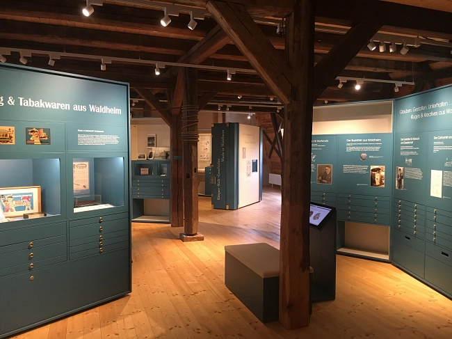 Das Bild zeigt einen Ausstellungsraum zur Stadtgeschichte von Waldheim. Der Raum befindet sich in einem historischen Gebäude mit sichtbaren Holzbalken und Holzboden. Mehrere große, türkisfarbene Ausstellungstafeln und -schränke stehen frei im Raum. Auf ihnen sind Texte, Fotos und Dokumente zur Geschichte von Waldheim zu sehen.
In der Mitte steht eine schmale, freistehende Stele mit weiteren Informationen. Daneben befindet sich eine Sitzbank für Besucherinnen und Besucher. Die Ausstellung ist modern gestaltet und gut ausgeleuchtet. Die Kombination aus historischen Holzbalken und modernen Ausstellungselementen schafft eine ruhige, übersichtliche Atmosphäre.