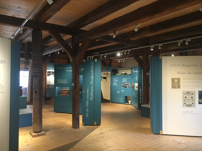 Das Bild zeigt einen weiteren Ausstellungsraum der Waldheimer Stadtgeschichte.
Der Raum befindet sich in einem historischen Gebäude mit sichtbaren, dunklen Holzbalken und einem hellen Holzboden. Mehrere türkisfarbene Ausstellungstafeln stehen frei im Raum und sind übersichtlich angeordnet. Auf den Tafeln sind Texte, Fotografien und historische Dokumente zu sehen.
Rechts im Vordergrund befindet sich eine helle Wandtafel mit einer Überschrift und einem historischen Porträt. Im Hintergrund führen die Ausstellungselemente weiter durch den Raum und geben den Blick auf zusätzliche Themenbereiche frei.
Die Beleuchtung erfolgt über Deckenspots und sorgt für eine gleichmäßige, angenehme Ausleuchtung. Die Kombination aus historischem Fachwerk und moderner Ausstellungsgestaltung schafft eine ruhige und strukturierte Atmosphäre.