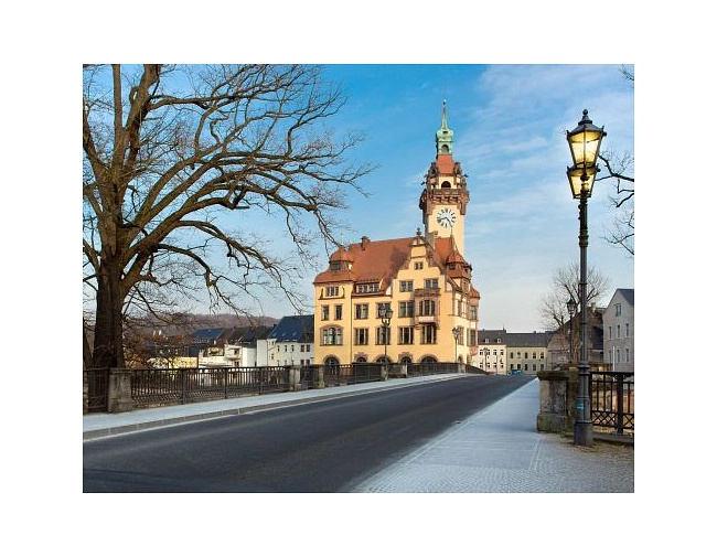 Das Bild zeigt das Rathaus von Waldheim.
Das Rathaus ist ein großes, historisches Gebäude mit mehreren Etagen. Die Fassade ist gelb gestrichen, das Dach ist rot. Besonders auffällig ist der hohe Turm mit einer Uhr und einer grünen Turmspitze.
Das Gebäude steht direkt an einer Brücke über einen Fluss. Die Straße auf der Brücke ist leer. Links sieht man einen großen, kahlen Baum. Rechts steht eine alte Straßenlaterne.

Im Hintergrund sind weitere Häuser und eine leicht hügelige Landschaft zu sehen. Der Himmel ist blau und klar. Das Bild wirkt ruhig und freundlich.