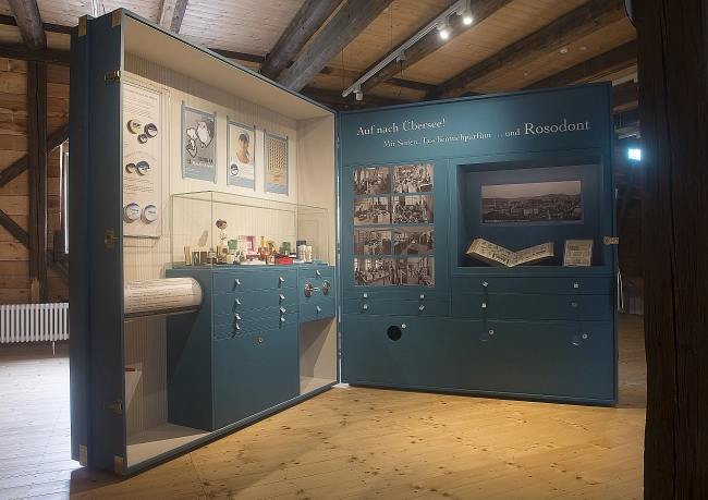 Das Bild zeigt einen modern gestalteten Ausstellungsraum zur Waldheimer Stadtgeschichte im Themenbereich „Florena“.
Der Raum befindet sich in einem historischen Gebäude mit sichtbaren dunklen Holzbalken an Decke und Wänden sowie einem hellen Holzboden. Mehrere große, türkisfarbene Ausstellungsmodule stehen frei im Raum. Sie enthalten gut lesbare Texte, historische Fotos und Dokumente zur Geschichte von Florena in Waldheim.
Im Hintergrund ist ein weiteres, freistehendes Ausstellungselement zu sehen. In der Mitte des Raumes steht eine schmale, interaktive Informationsstele. Eine schlichte Sitzbank lädt zum Verweilen ein.
Die Ausstellung ist hell und gleichmäßig ausgeleuchtet. Die Kombination aus historischen Bauelementen und moderner Präsentation schafft eine übersichtliche und ruhige Atmosphäre.