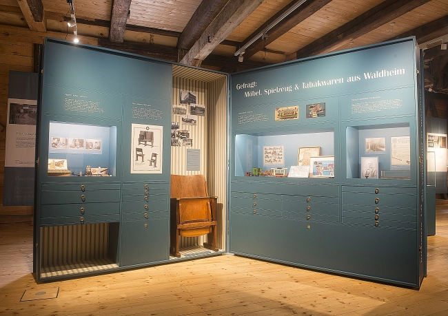 Das Bild zeigt einen Ausstellungsbereich zur Industrie- und Wirtschaftsgeschichte von Waldheim.
Im Mittelpunkt stehen zwei große, türkisfarbene Ausstellungstafeln, die im rechten Winkel zueinander aufgestellt sind. Auf ihnen befinden sich Texte, historische Fotografien, Zeichnungen und Dokumente.
Unterhalb der Tafeln sind mehrere flache Schubladen integriert. Zwischen den beiden Tafeln ist ein historischer Friseurstuhl aus Holz mit gepolsterter Sitzfläche ausgestellt.
Der Raum befindet sich in einem historischen Gebäude mit sichtbaren Holzbalken und einem hellen Holzboden. Die Beleuchtung durch Deckenspots sorgt für eine gleichmäßige Ausleuchtung. Die Präsentation verbindet historische Architektur mit moderner Ausstellungsgestaltung und wirkt übersichtlich und ruhig.