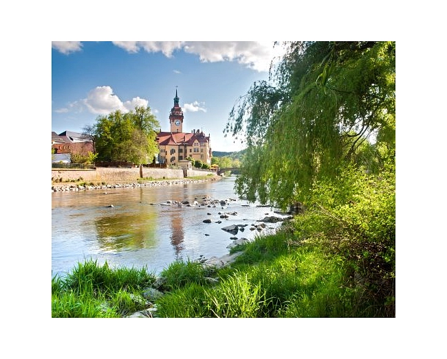 Das Bild zeigt das Rathaus von Waldheim an der Zschopau.
Das Rathaus ist ein großes, historisches Gebäude mit gelber Fassade und rotem Dach. In der Mitte steht ein hoher Turm mit einer Uhr und einer dunklen Turmspitze. Das Gebäude liegt direkt am Ufer.
Im Vordergrund sieht man den Fluss. Das Wasser ist ruhig, und einige Steine ragen aus dem Wasser heraus. Am Ufer wächst grünes Gras. Rechts im Bild hängt ein großer Baum mit langen, grünen Zweigen über das Wasser.
Der Himmel ist blau mit weißen Wolken. Das Sonnenlicht lässt das Rathaus und die Natur hell und freundlich wirken.
Das Bild zeigt eine ruhige, idyllische Landschaft mit dem Rathaus von Waldheim im Mittelpunkt.