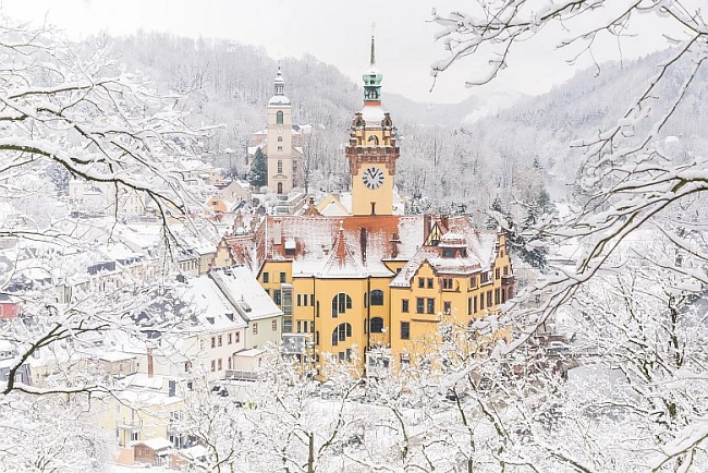 Das Bild zeigt eine winterliche Ansicht der Stadt Waldheim. Im Vordergrund sind kahle, mit Schnee bedeckte Äste von Bäumen zu sehen, die einen Rahmen um die Szene bilden. In der Mitte des Bildes steht das Rathaus von Waldheim, ein historisches Gebäude mit einem markanten Uhrturm und einer auffälligen gelben Fassade. Der Schnee bedeckt das gesamte Dach sowie die umliegenden Dächer und Landschaften. Im Hintergrund sind weitere Gebäude sichtbar, darunter die Kirche, die mit ihrem Turm ebenfalls sehr prägnant in der winterlichen Szenerie hervorsticht. Die ruhige Atmosphäre des Bildes spiegelt das malerische Stadtbild wider, das durch die Architektur der beiden Gebäude – das Rathaus und die Kirche – dominiert wird. Das Bild hat einen fast märchenhaften Charakter, der durch den klaren Himmel und die schneebedeckten Bäume noch verstärkt wird.