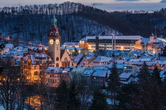 Winterliche Abendaufnahme der Stadt Waldheim. Im Vordergrund stehen dicht beieinanderliegende Häuser mit schneebedeckten Dächern. Warmes Licht scheint aus Fenstern und Straßenlaternen und taucht die Altstadt in eine ruhige Atmosphäre. Links im Zentrum ragt das Rathaus mit seinem hohen Turm und der beleuchteten Uhr deutlich sichtbar über die Dächer hinaus.

Im Hintergrund erstreckt sich ein großes, langgezogenes Gebäude auf einer Anhöhe – die Justizvollzugsanstalt (JVA). Dahinter liegt ein bewaldeter, leicht verschneiter Hang. Der Himmel ist in der Dämmerung blau-grau gefärbt. Die Gesamtstimmung wirkt ruhig und winterlich.