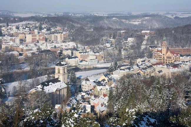Das Bild zeigt die Stadt Waldheim in Sachsen im Winter, aufgenommen vom Wachbergturm aus.

Im Vordergrund sind verschneite Bäume zu sehen. Dahinter liegt die Altstadt mit vielen dicht stehenden Häusern. Die Dächer sind mit Schnee bedeckt. Die Gebäude haben helle Fassaden in Weiß, Beige und Pastelltönen.

Links im Bild steht eine Kirche mit einem dunklen Zwiebelturm. Rechts ist das Rathaus mit einem hohen, rot verklinkerten Turm zu sehen. Der Turm hebt sich deutlich von den umliegenden Gebäuden ab.

In der Mitte fließt die Zschopau durch die Stadt. Der Fluss ist nicht zugefroren und schlängelt sich zwischen den Häusern hindurch.

Im Hintergrund erstrecken sich weitere Wohnhäuser, bewaldete Hügel und eine verschneite Landschaft. Der Himmel wirkt hell und leicht diesig. Das Bild vermittelt eine ruhige, winterliche Atmosphäre mit weitem Blick über Waldheim.