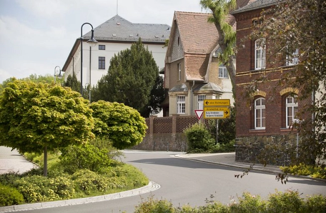 Die Aufnahme zeigt die Schlossstraße in Waldheim. Eine ruhige, leicht geschwungene Straße führt durch ein gepflegtes Wohngebiet. Im Vordergrund befindet sich eine begrünte Verkehrsinsel mit niedrigen Sträuchern und einem kleinen, rund geschnittenen Baum.
Entlang der Straße stehen historische Gebäude mit unterschiedlichen Fassaden. Rechts ist ein mehrgeschossiges Backsteingebäude mit weißen Fensterrahmen zu sehen. Daneben stehen weitere Häuser mit hell verputzten Wänden und geneigten Dächern.
An der Straßenecke befindet sich ein gelbes Wegweiserschild. Straßenlampen säumen den Gehweg. Die Umgebung wirkt sauber, ruhig und von viel Grün geprägt.