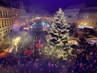 Weihnachtsmarkt Waldheim_BF Das Bild zeigt eine festliche Szene von einem Weihnachtsmarkt bei Nacht. Im Vordergrund ist ein üppig geschmückter Weihnachtsbaum zu sehen, der mit vielen Lichtern erleuchtet ist. Der Baum steht vor einer großen Menschenmenge, die sich durch die Straßen des Marktes bewegt. Es gibt viele Stände, die bunte Lichter und Deko aufweisen, und einige der Stände verkaufen vermutlich Waren oder Speisen. Im Hintergrund sieht man die Fassaden von Gebäuden, die ebenfalls in weihnachtlicher Beleuchtung erstrahlen. Der Himmel ist dunkel und es scheint leicht neblig, was die festliche Atmosphäre verstärkt. Die Menge ist gut sichtbar und es gibt eine bunte Mischung von Menschen, die warme Kleidung tragen, was darauf hinweist, dass es kalt ist. © Grit Lässig